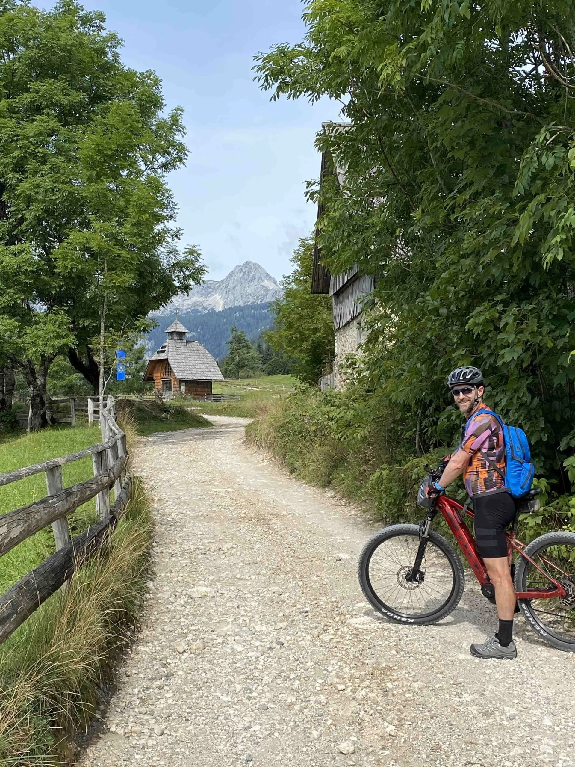 Mountain biker with red bike on gravel path near wooden cabin and mountains