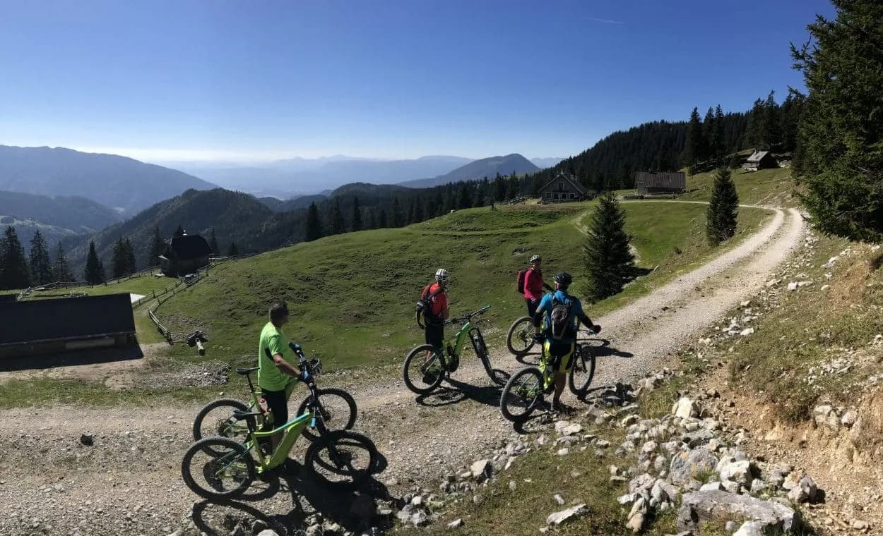 Mountain bikers on a gravel path overlooking rolling green hills and distant mountains under a clear blue sky.