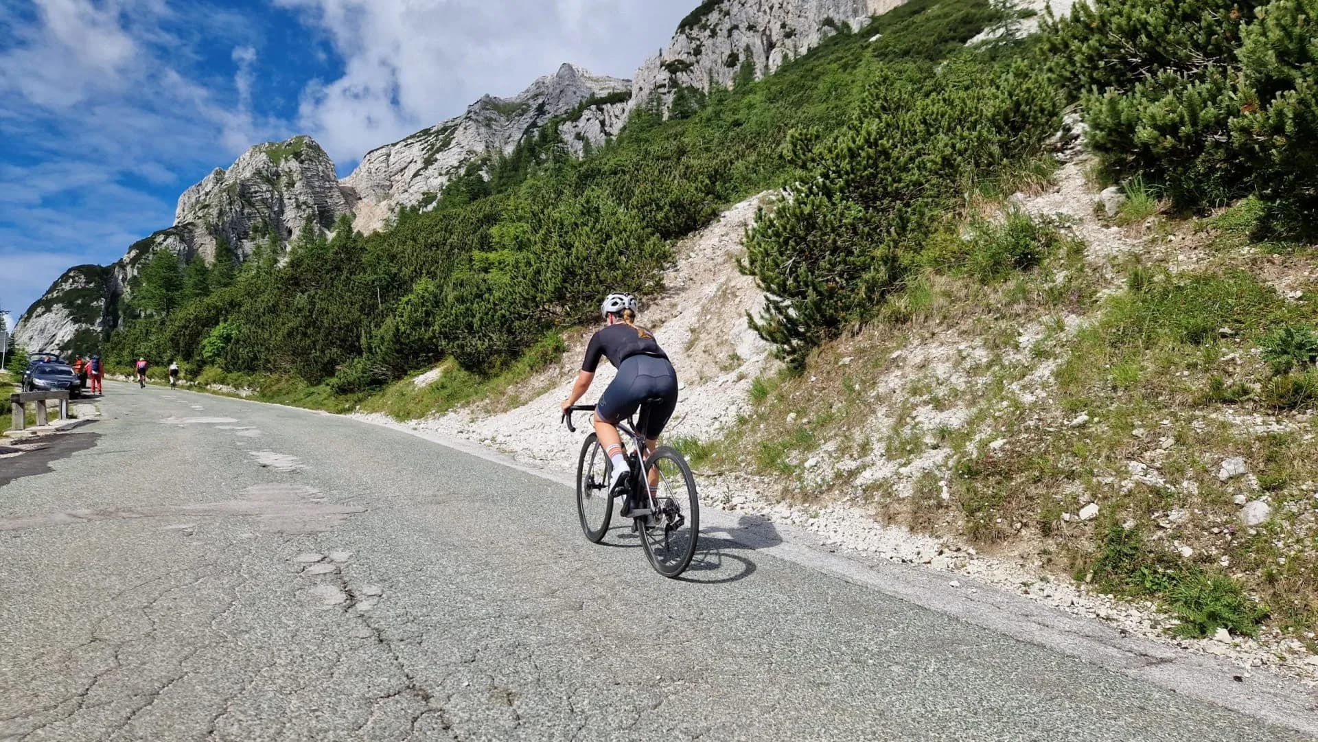 Cyclist riding up cracked asphalt road alongside steep, rocky mountainside with green shrubs.