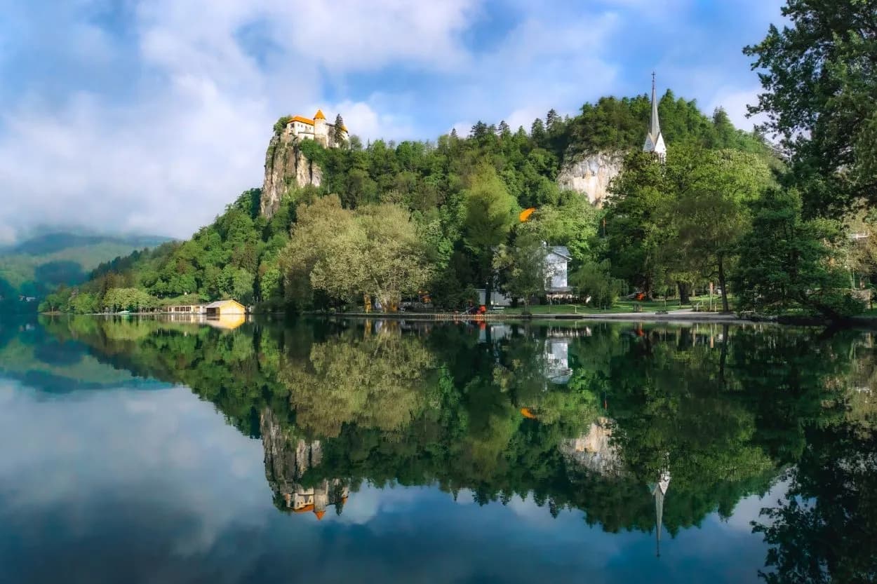 Bled Castle and church spire reflected in calm Lake Bled surrounded by green forest and hills.