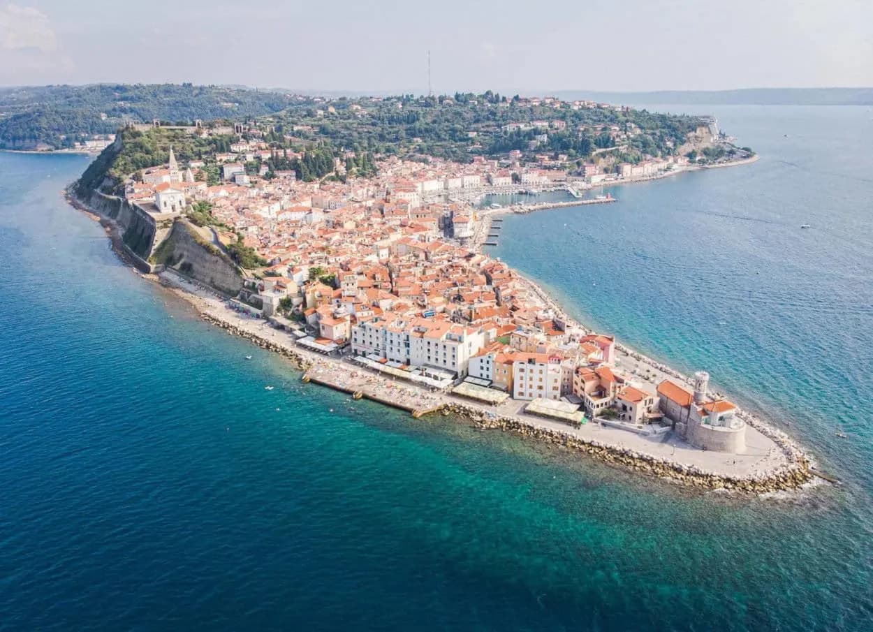 Aerial view of Piran peninsula town with red roofs surrounded by turquoise Adriatic Sea water.