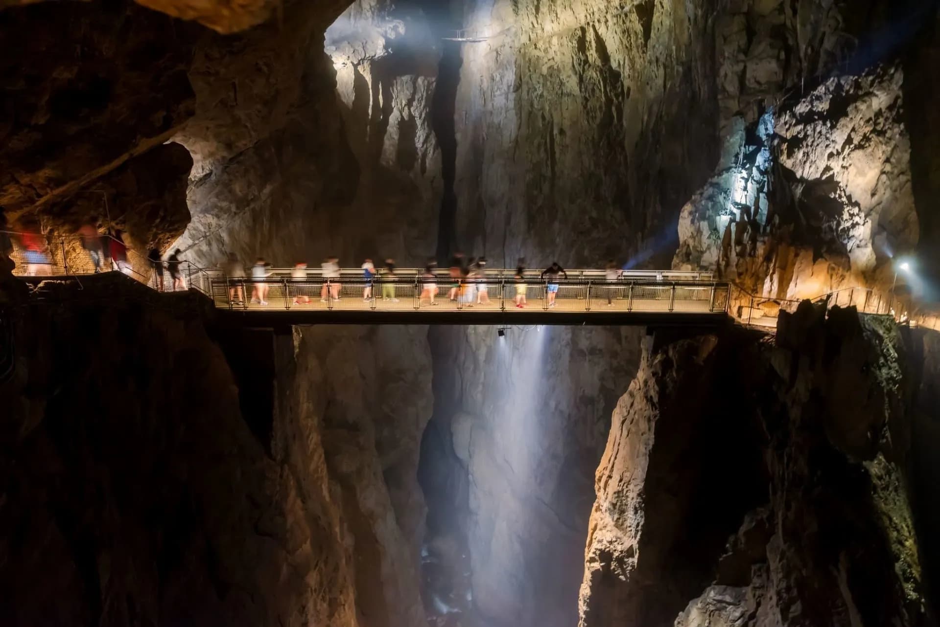 Tourists crossing a lighted bridge over a deep chasm inside Škocjan Caves, Slovenia.