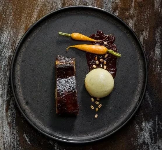 Gourmet plate with glazed meat, baby carrots, pale green sphere, and berry reduction.