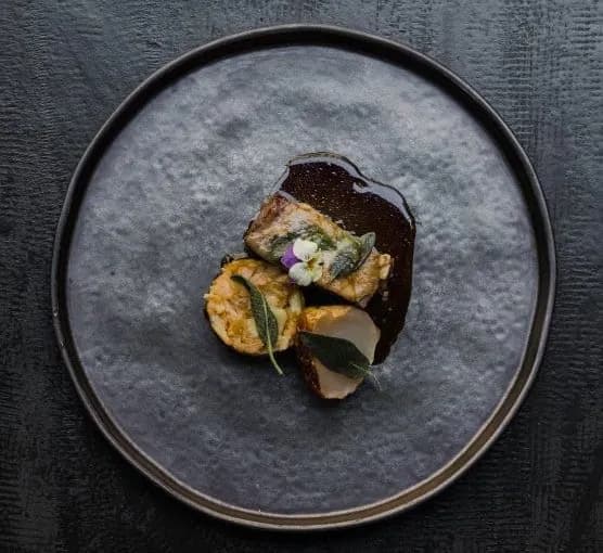 Gourmet plated food with meat, vegetables, dark sauce, and edible flowers on a dark plate.