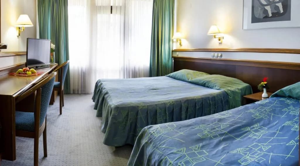 Standard hotel room with two beds, a desk, and bright window light.