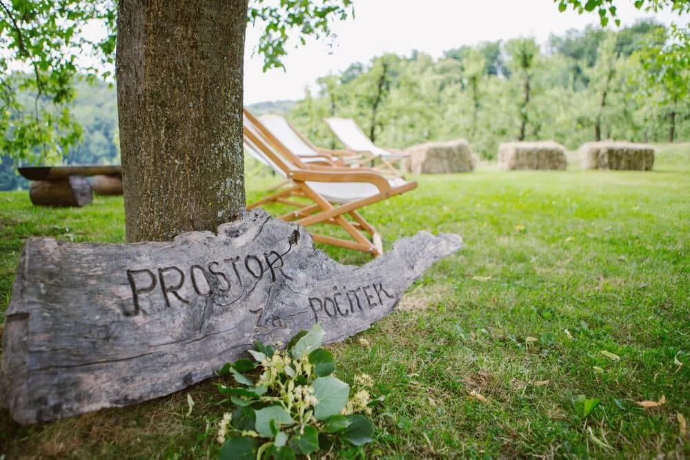 Wooden sign reading "Prostor za počitek" by tree with deck chairs in a grassy garden.