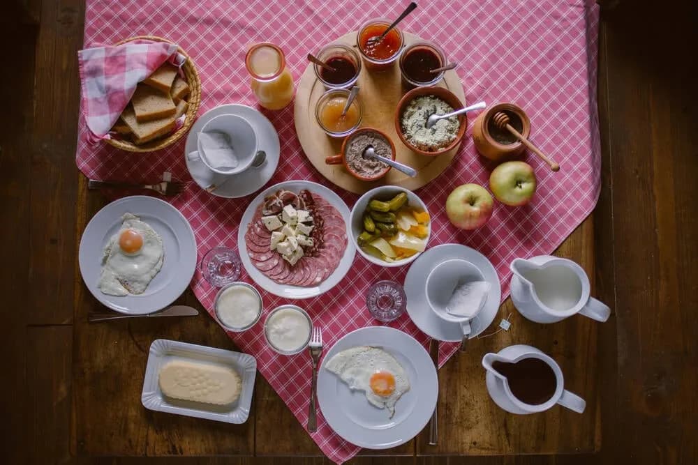 Hearty breakfast spread with fried eggs, cured meats, cheese, bread, jams, and apples on a wooden table.