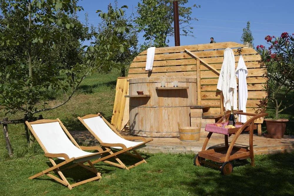 Outdoor wooden hot tub area with lounge chairs, robes, and a drinks cart in a grassy setting.