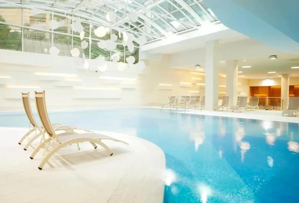Indoor swimming pool with glass roof, white modern design, and lounge chairs at Hotel Mirna.