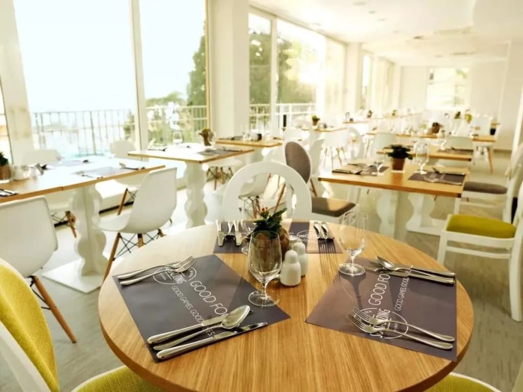 Bright breakfast area with wooden tables, white chairs, and large windows at Hotel Mirna