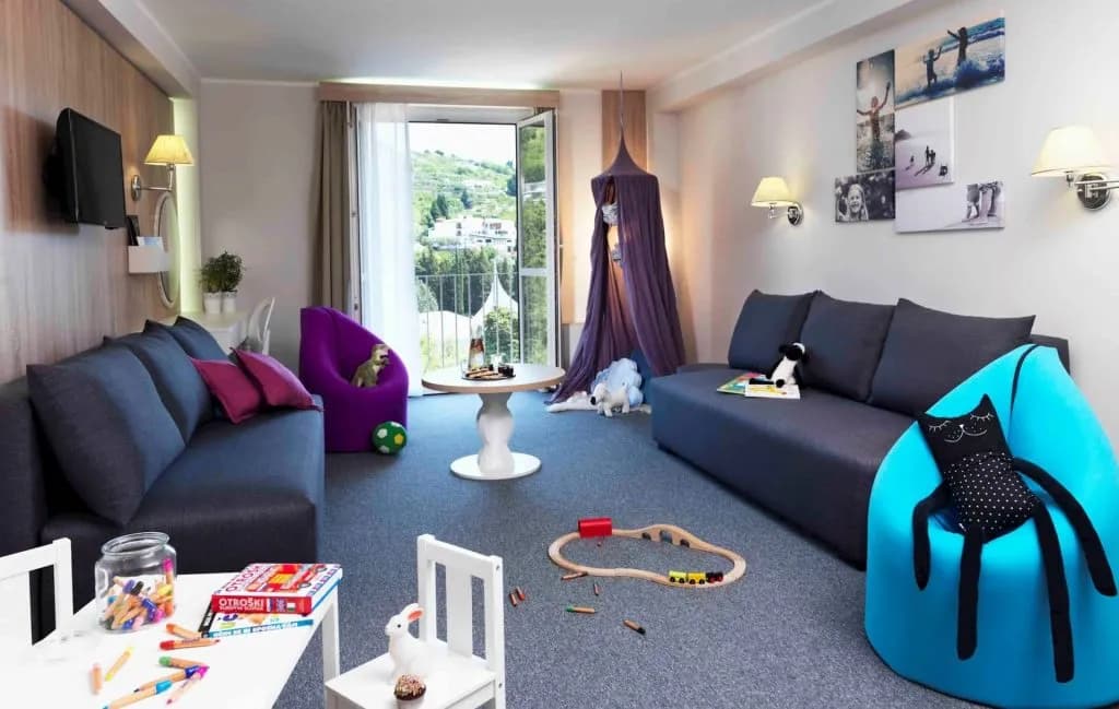 Hotel Mirna children's room with sofas, toys, and balcony view of green hillsides.