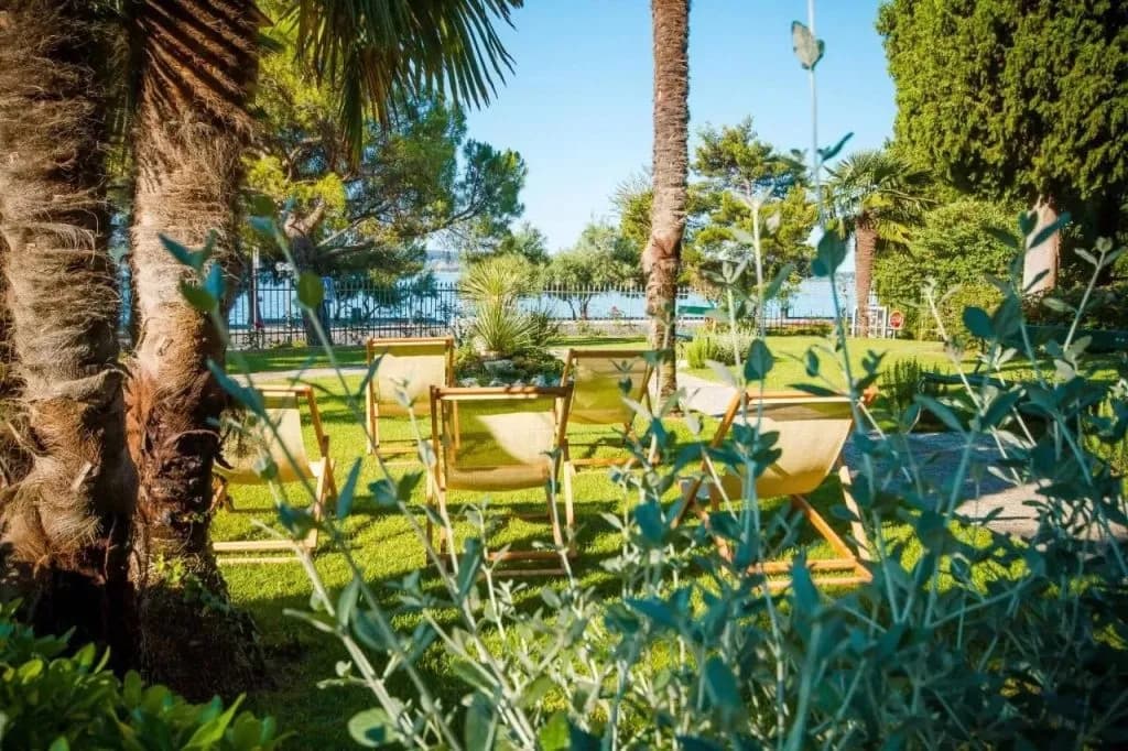 Lawn chairs in a sunny garden with palm trees overlooking the sea at Hotel Marko.