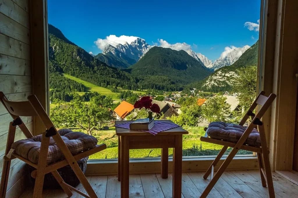 Mountain view from wooden cabin balcony with chairs, table, and roses overlooking Dovje village.