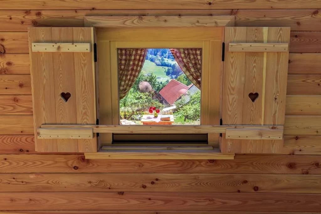 Wooden window with open shutters showing a green mountain view from a log cabin in Dovje.