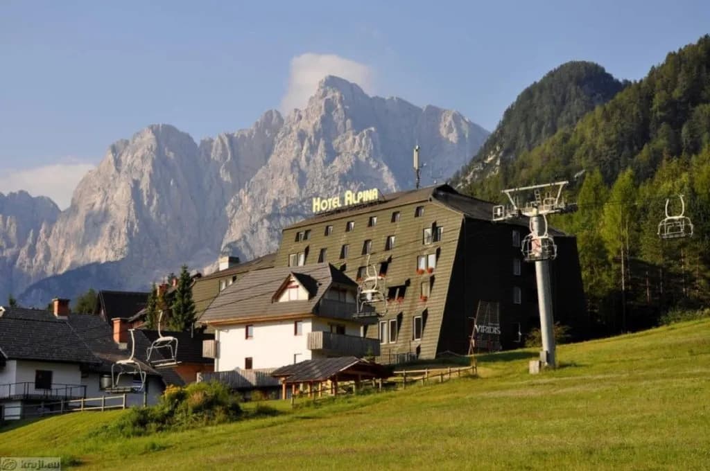 Hotel Alpina building with ski lift chairs in front of rugged mountains and green slopes.