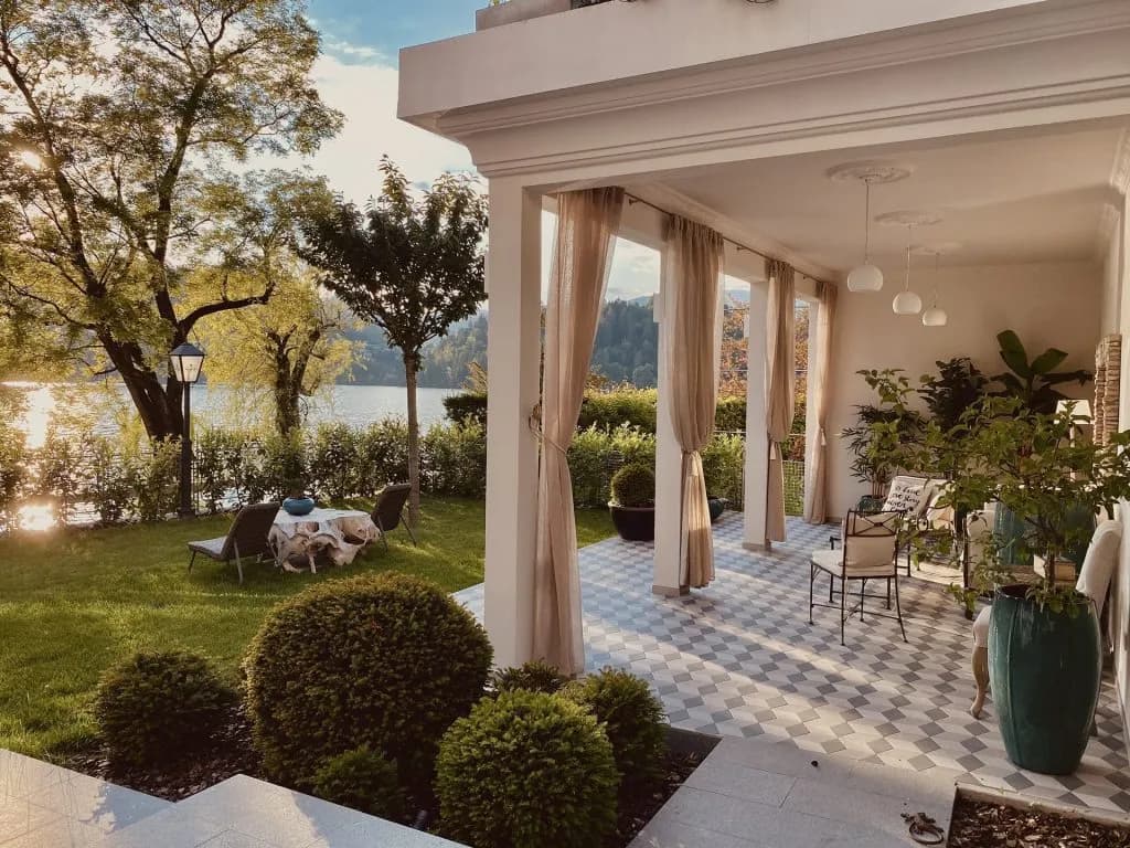 Covered patio overlooking a lake with manicured lawn, trees, and sunlit water.