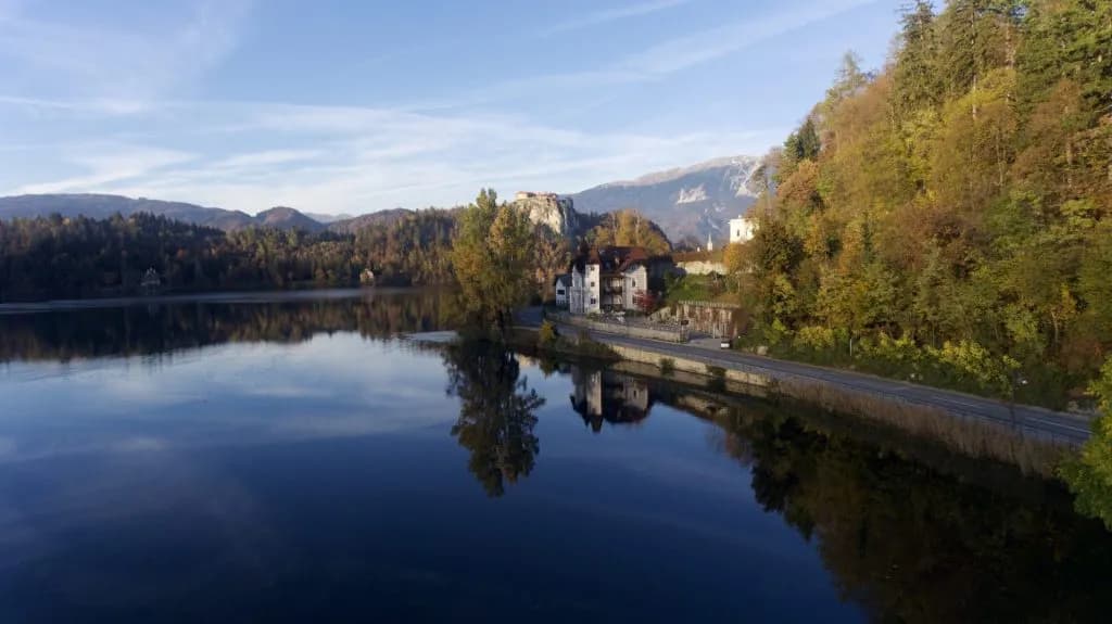 Calm lake reflecting autumn trees, buildings, and mountains near a road.