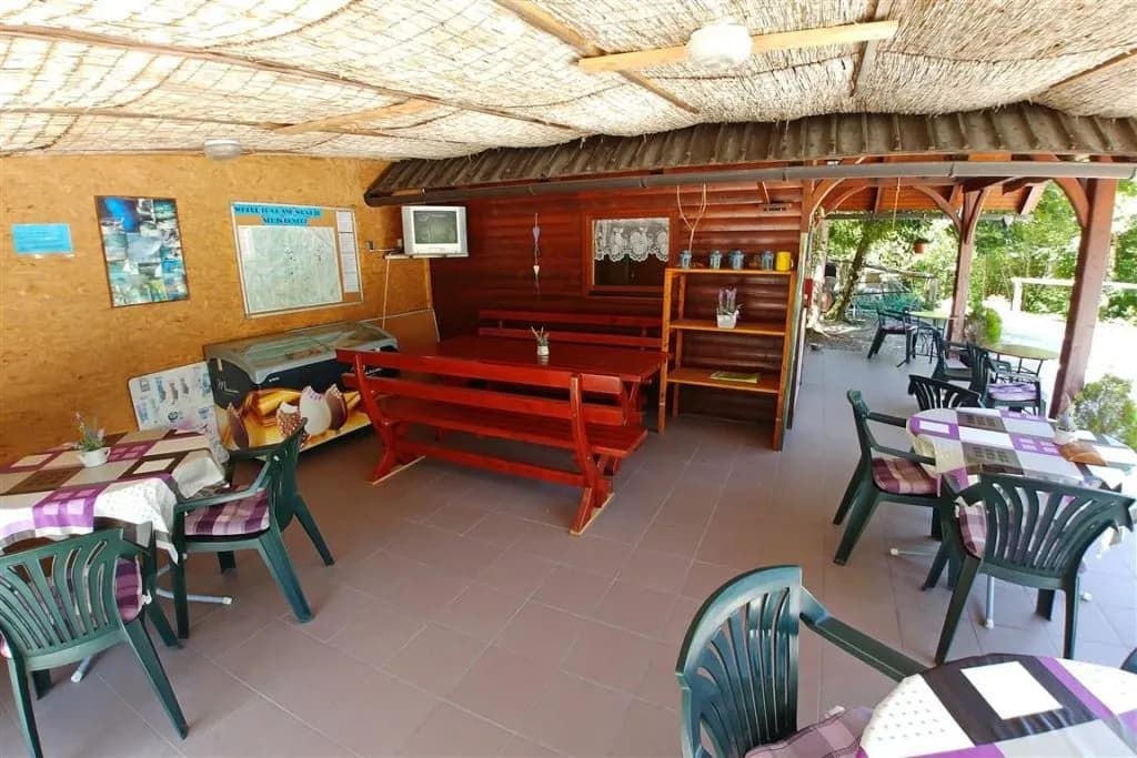 Covered outdoor bar area with wooden benches, plastic chairs, and a thatched roof near Vodenca.
