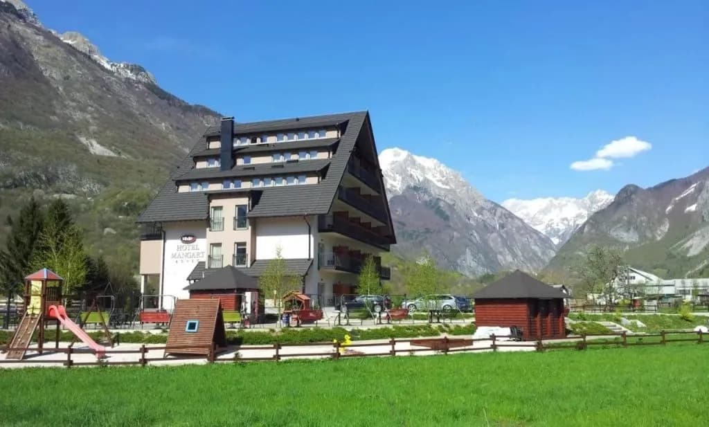 Hotel Mangart with playground in front of snow-capped mountains under blue sky.