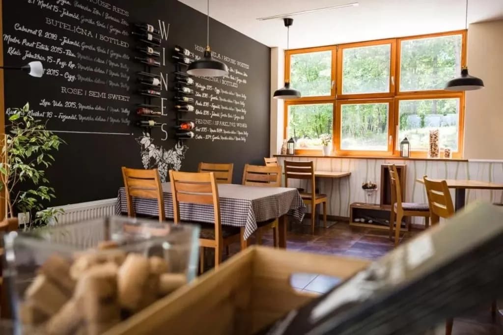 Dining area with wooden chairs, wine menu chalkboard, and large windows showing greenery.