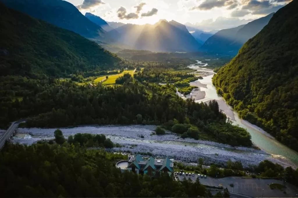 Aerial view of hotel by wide river valley, dense forest, and sun rays over mountains