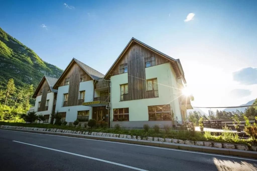 Hotel building with wood accents next to a road, set against a steep green mountain under bright sun.