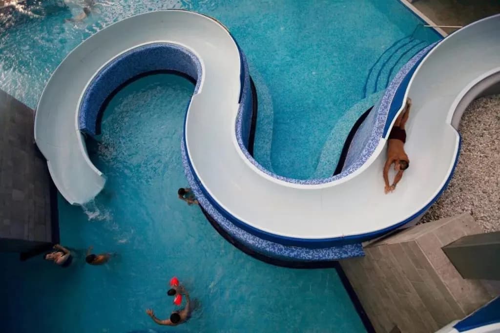 Man sliding down a white water slide into a blue indoor pool with other swimmers.