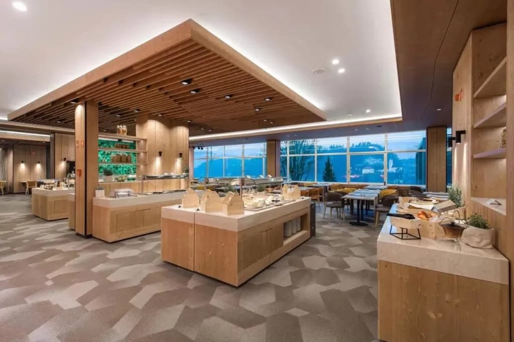 Hotel breakfast area with wood accents, buffet stations, and large windows overlooking a town.