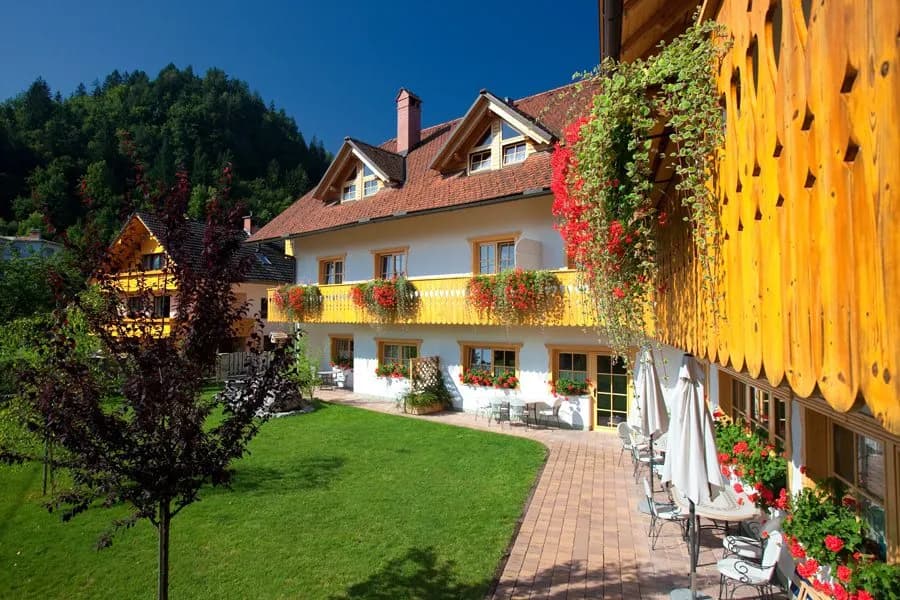 Garni hotel with white facade, yellow balconies, and red geraniums set against a green forested hill.
