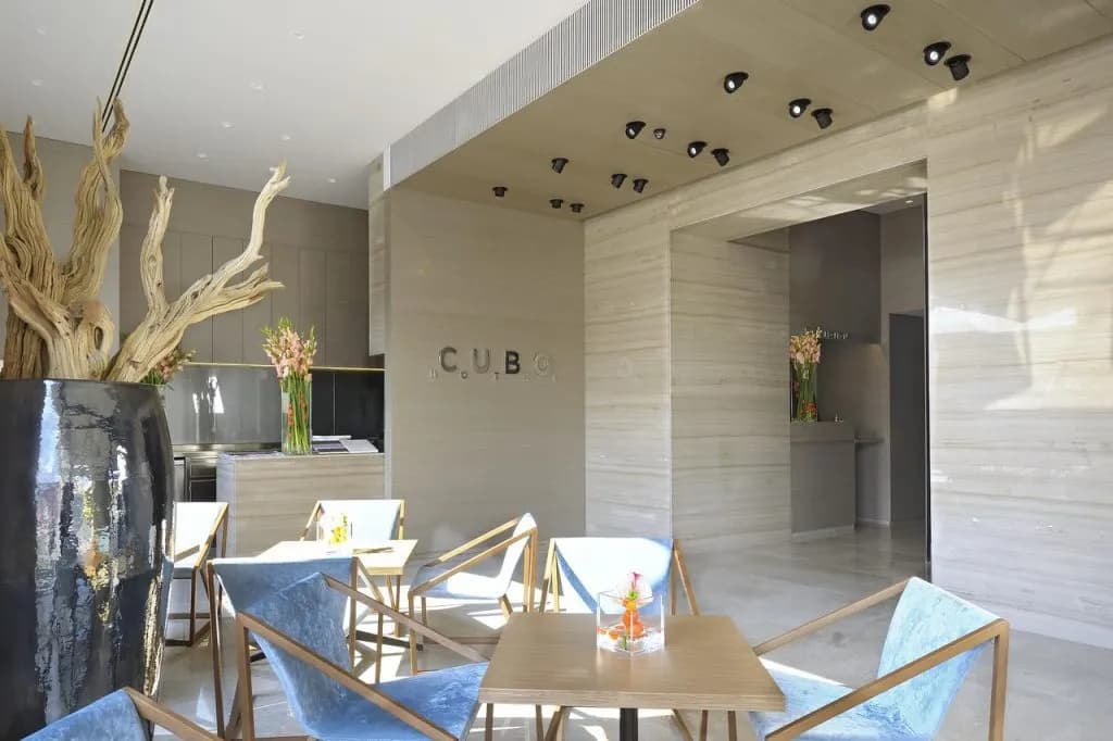 Modern hotel lobby seating area with light wood tables and blue velvet chairs, featuring the CUBO logo.