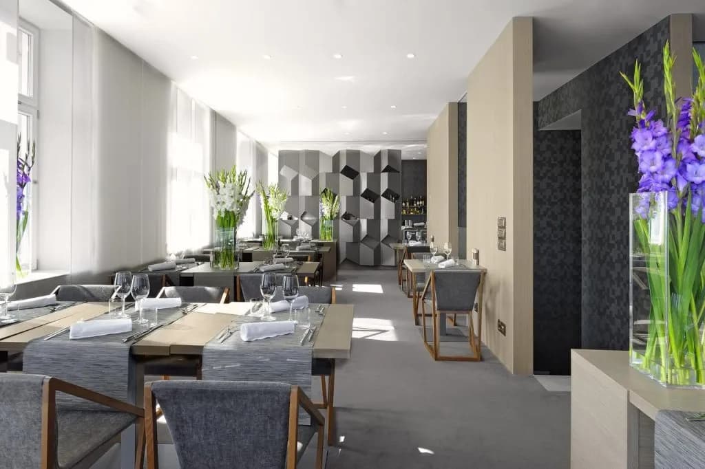 Modern hotel breakfast area with set tables, gray chairs, and tall flower arrangements.