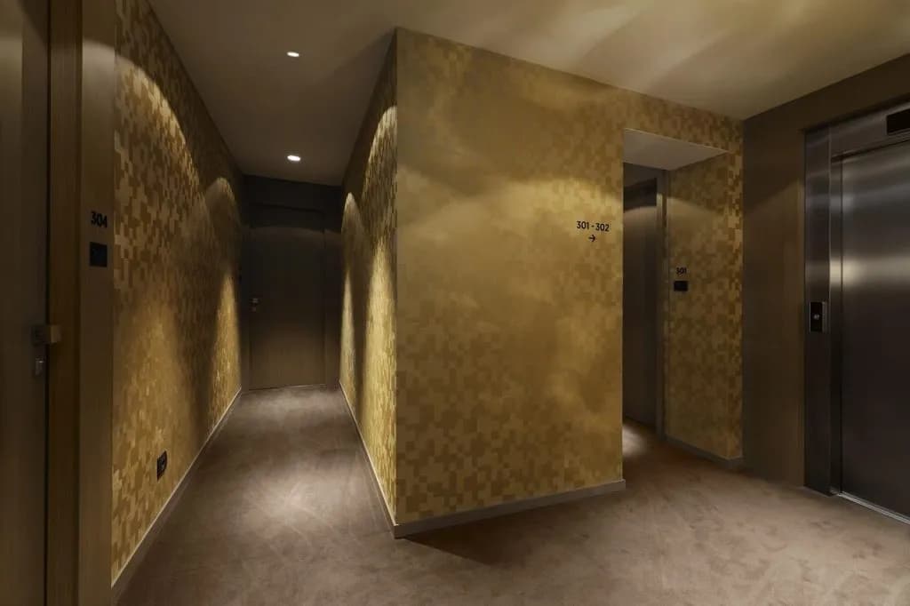 Hotel hallway interior with gold textured walls, recessed lighting, and stainless steel elevators.