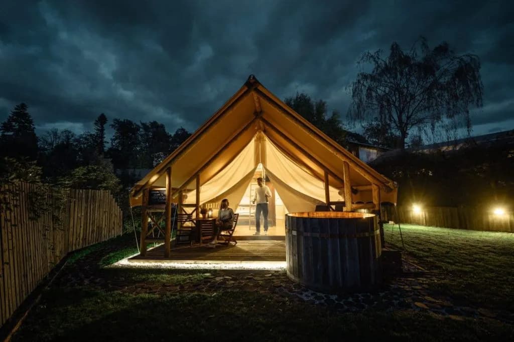 Illuminated glamping tent at night with wooden hot tub and two people near Bled.