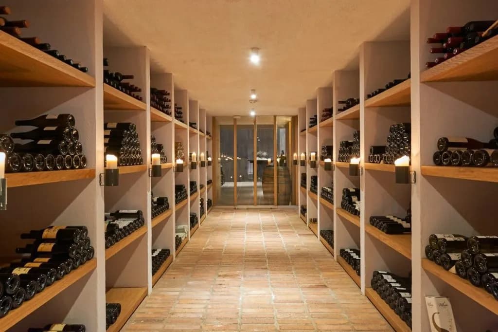 Wine cellar aisle with stacked bottles on wooden shelves illuminated by wall sconce candles and a barrel visible.