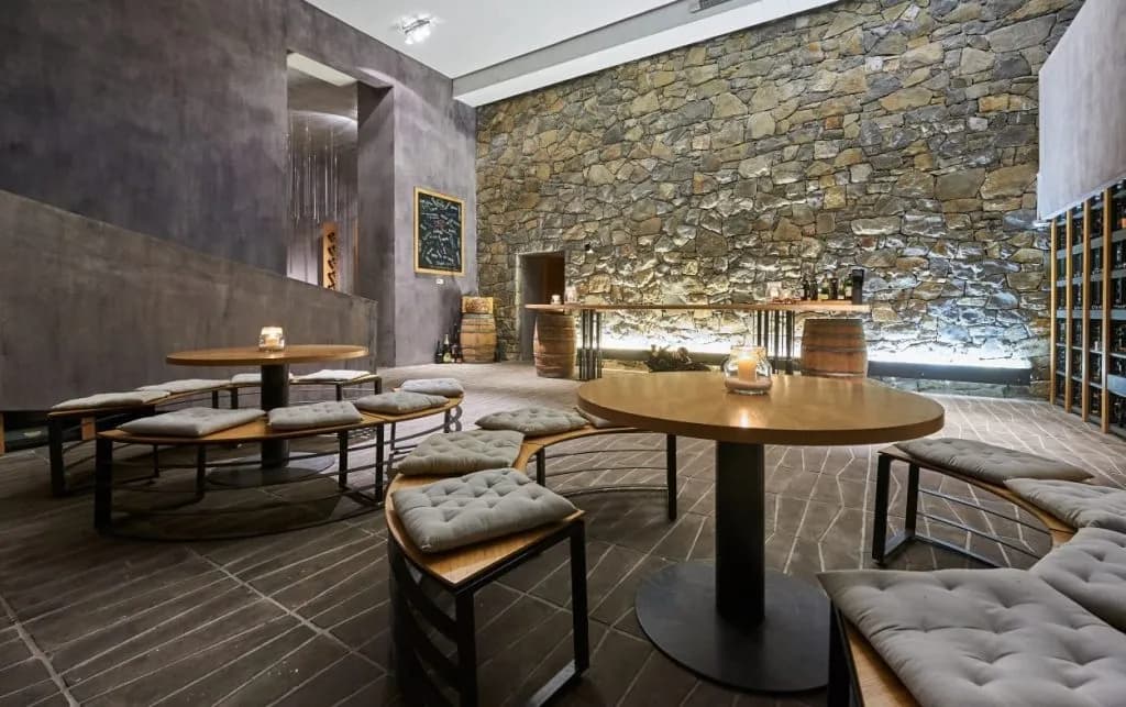 Wine tasting room with stone wall, wooden tables, and curved benches with cushions.