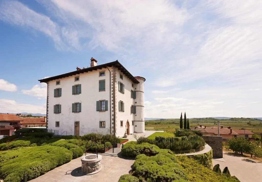 White historic building with green shutters and stone well in sunny landscaped garden
