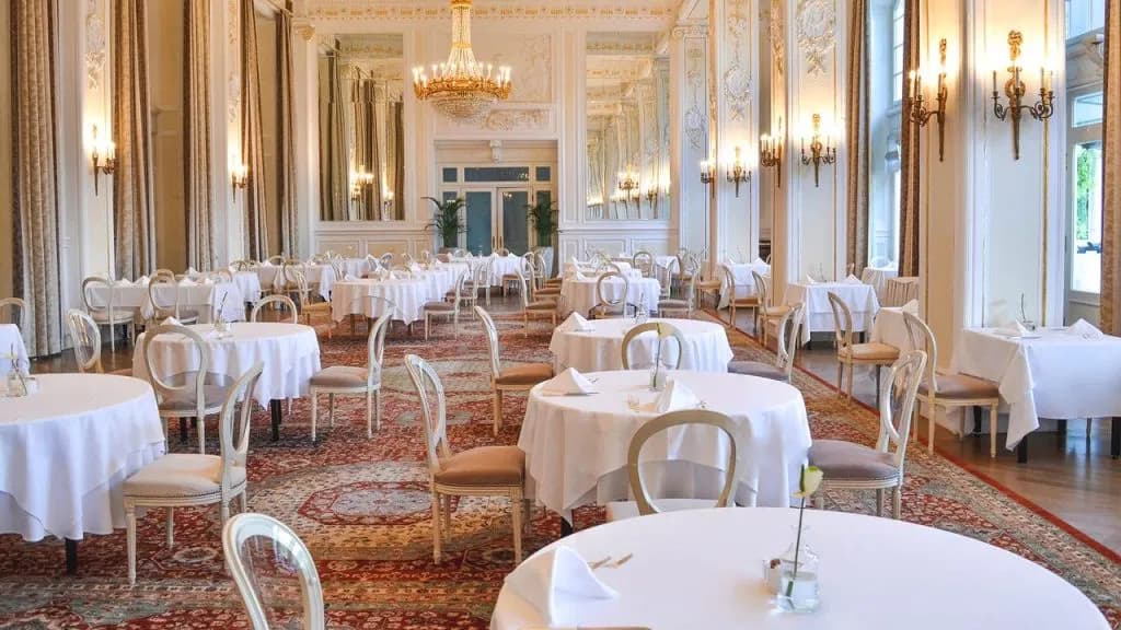 Elegant dining room with white linen tables, ornate chairs, and a large chandelier at Hotel Kempinski Palace.