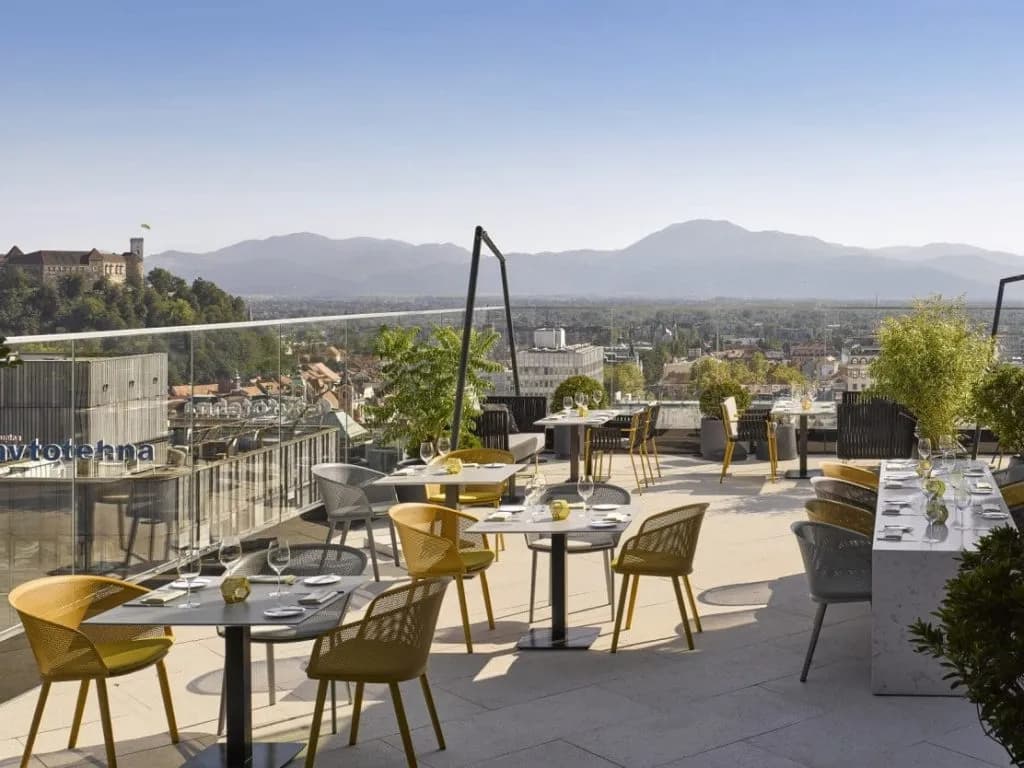 Rooftop restaurant terrace with tables set, overlooking Ljubljana Castle and mountains.