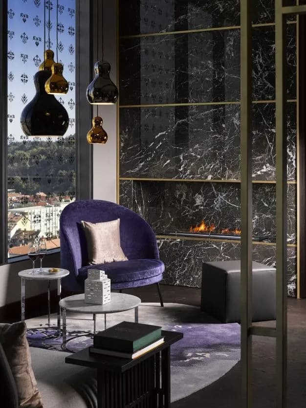 Luxury lounge with purple velvet chair, black marble fireplace, and hanging pendant lights overlooking a city.