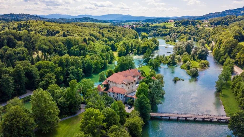 Aerial view of Grad Otočec castle surrounded by lush forest and a river with a bridge.