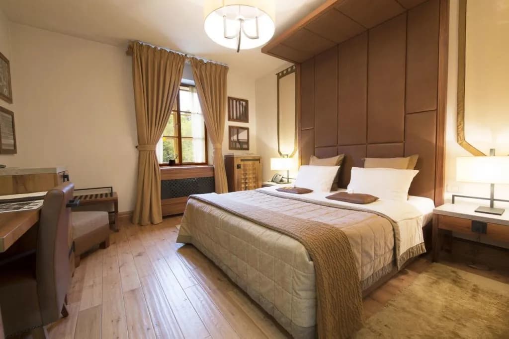Hotel room with large bed, brown headboard, light wood floor, and window with tan curtains.