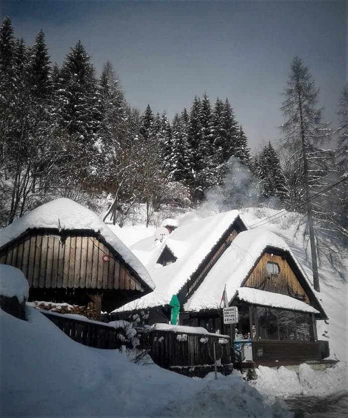 Wooden mountain huts with thick snow on roofs, smoke rising, surrounded by snow-covered pine forest.