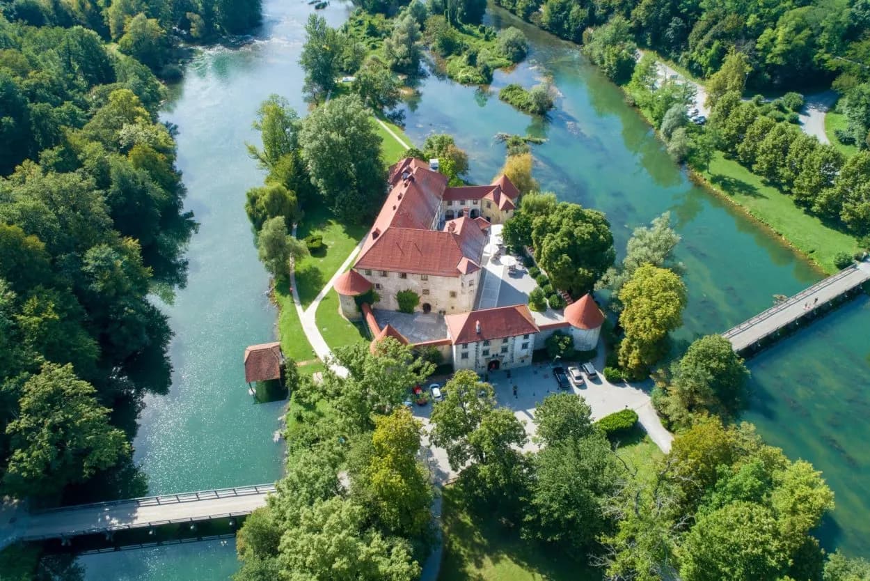 Otočec Castle surrounded by lush green trees and turquoise river water with two bridges.