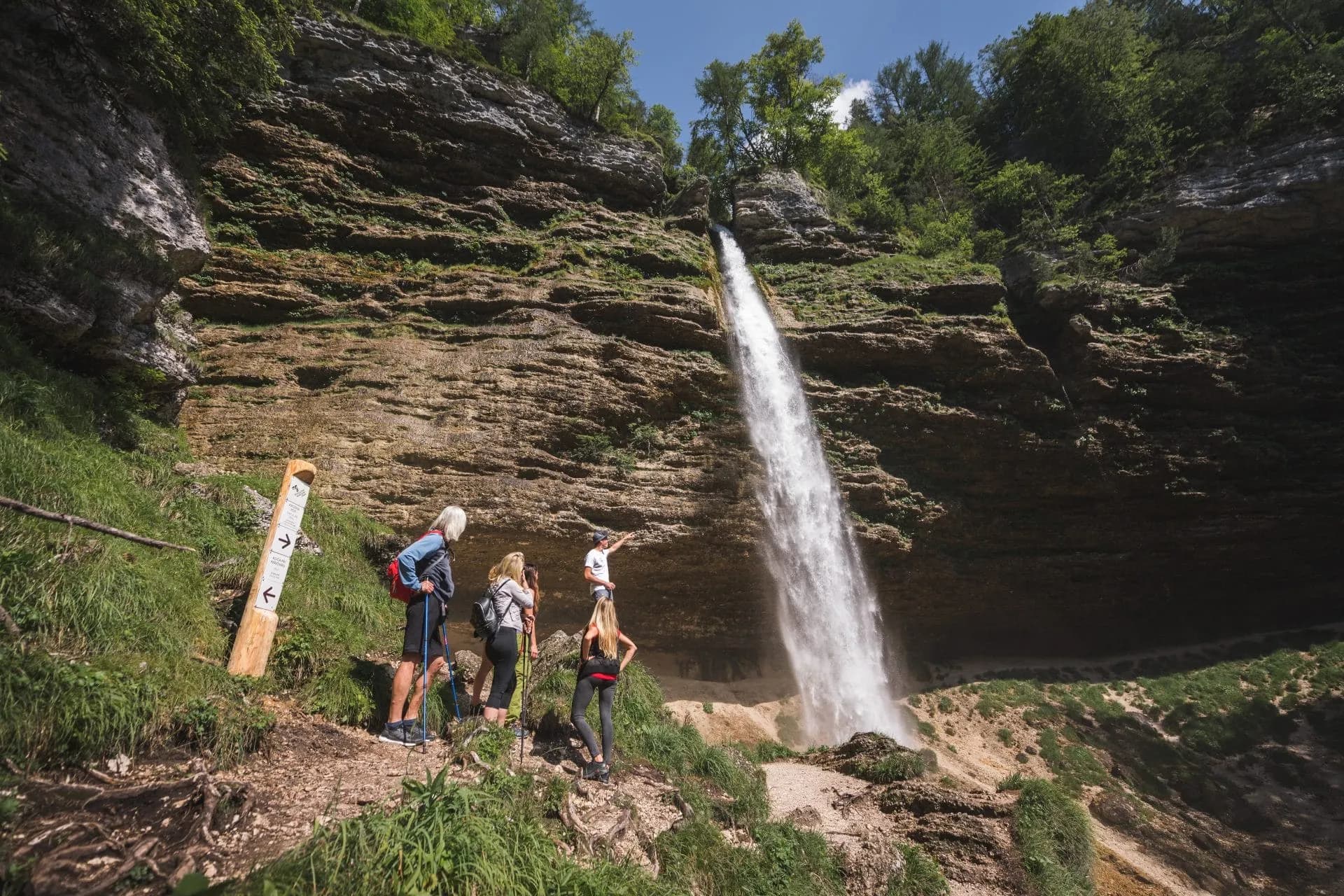 Hikers viewing Pericnik Waterfall cascading down a steep, rocky cliff face in summer.