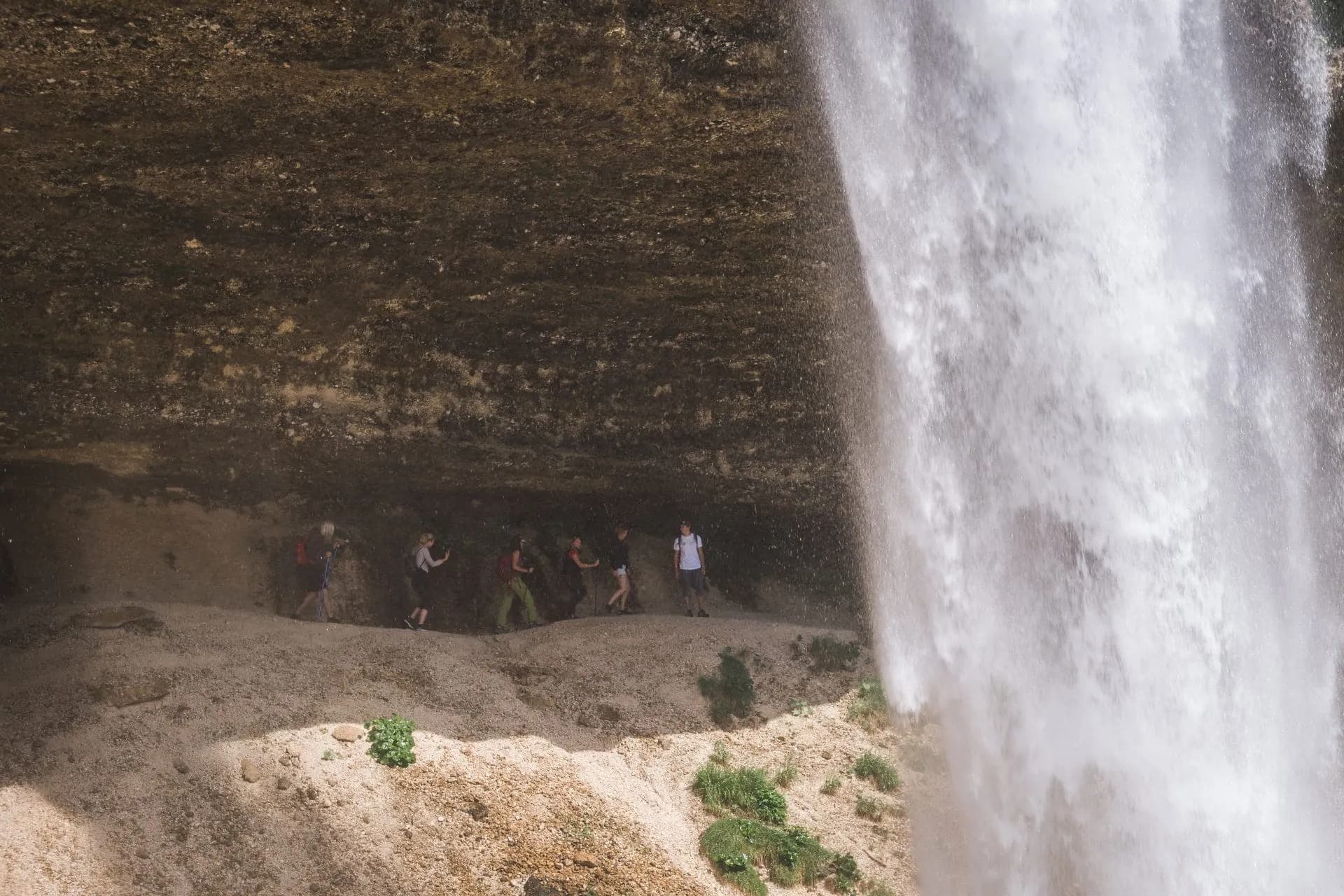Hikers walking behind the curtain of Pericnik Waterfall in a rocky alcove