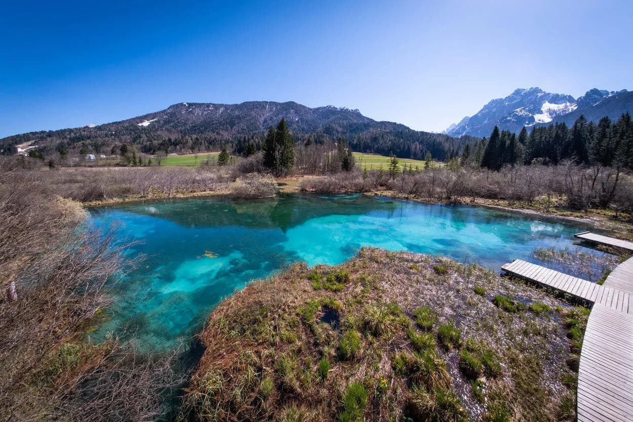 Turquoise spring water at Zelenci with wooden boardwalk and snow-capped Julian Alps in background.