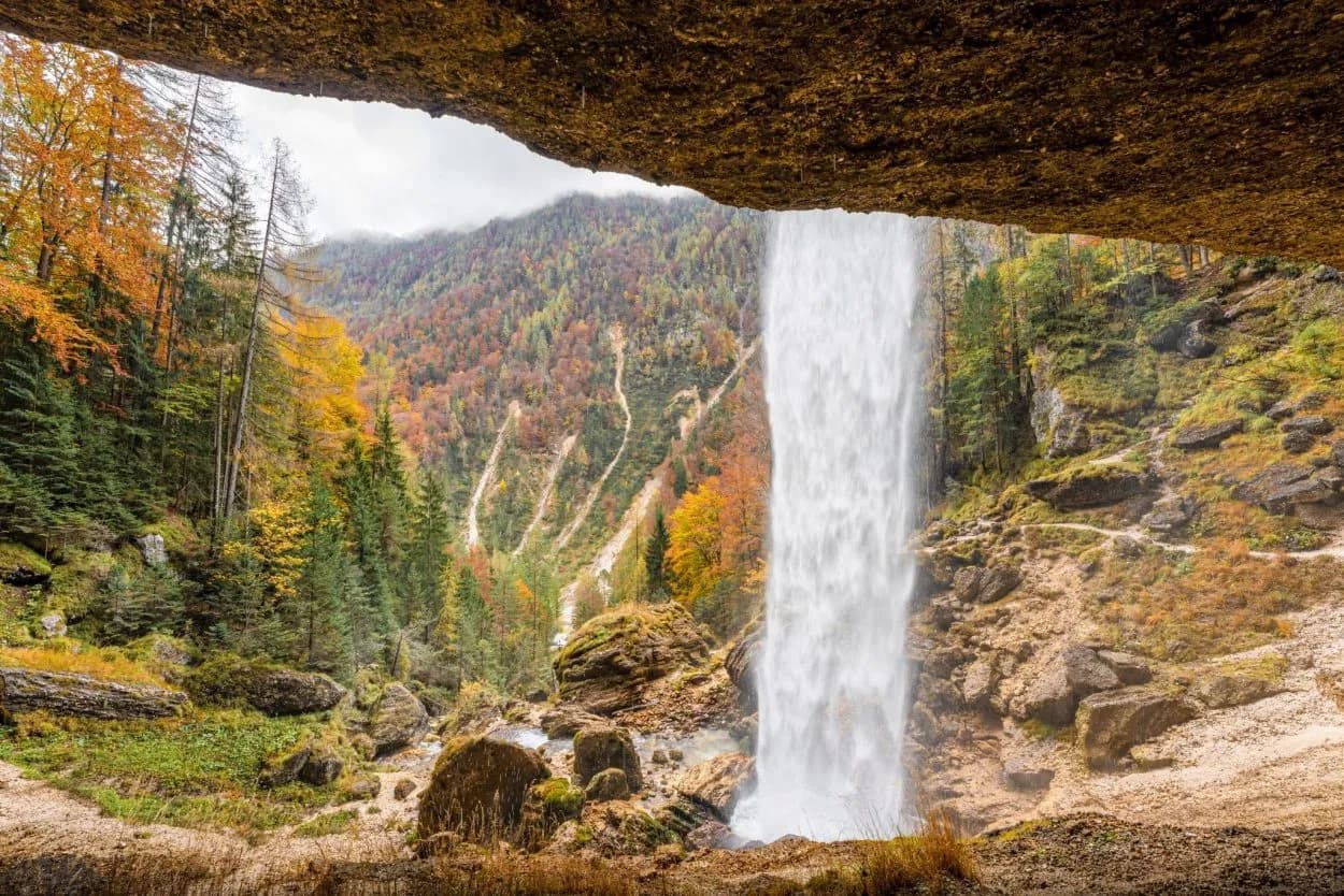 Waterfall plunging from behind an overhang into a rocky area with autumn forest on the slopes.