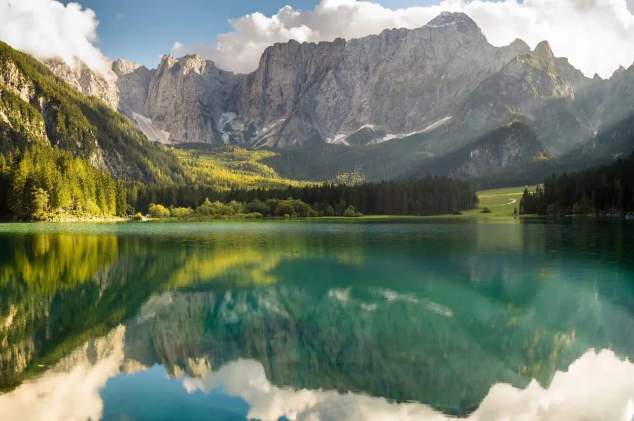 Mountain lake with turquoise water reflecting rugged peaks and green forests under a partly cloudy sky.