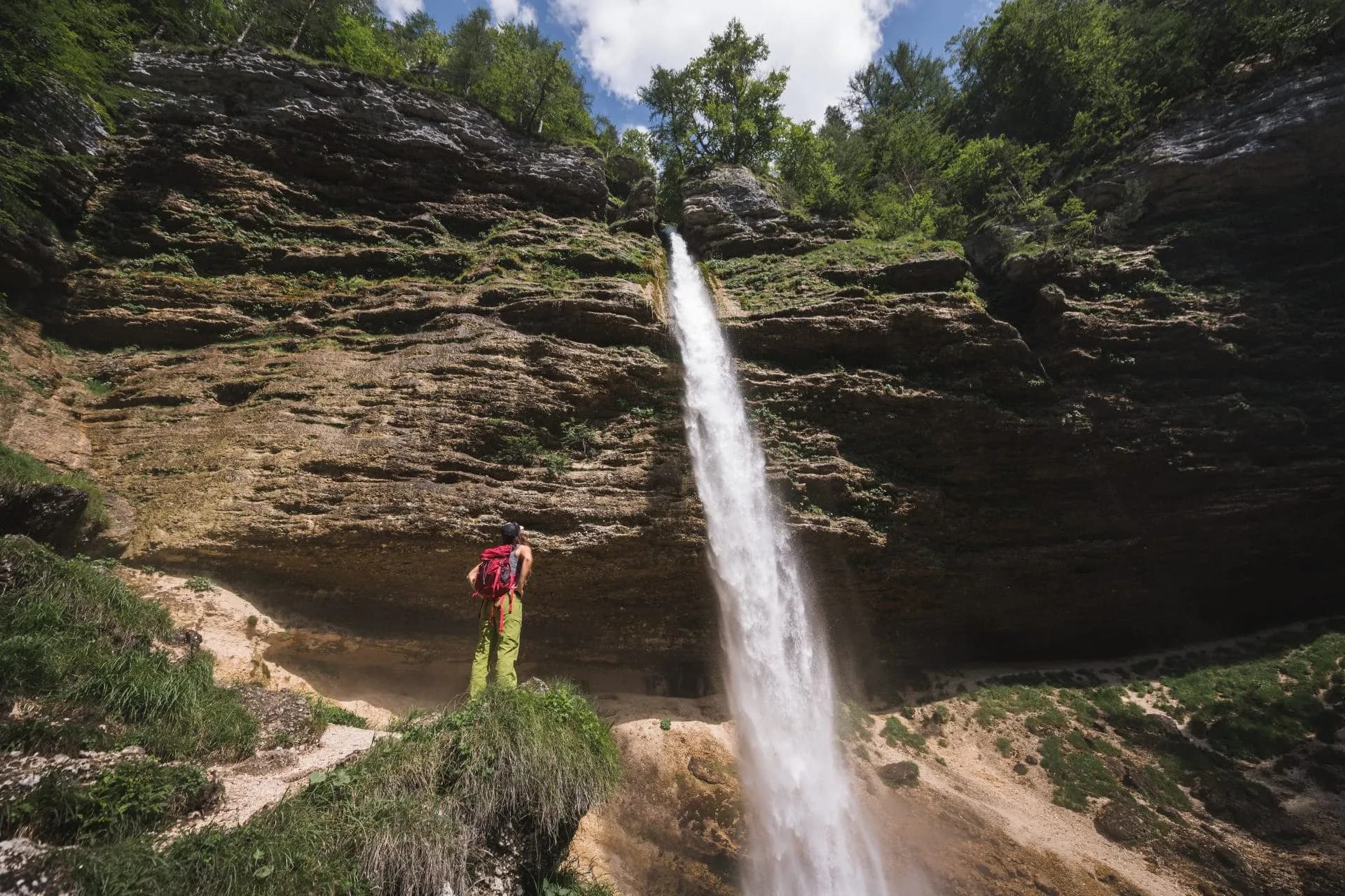 Hiker with red backpack observing tall waterfall cascading down mossy cliff face at Pericnik.