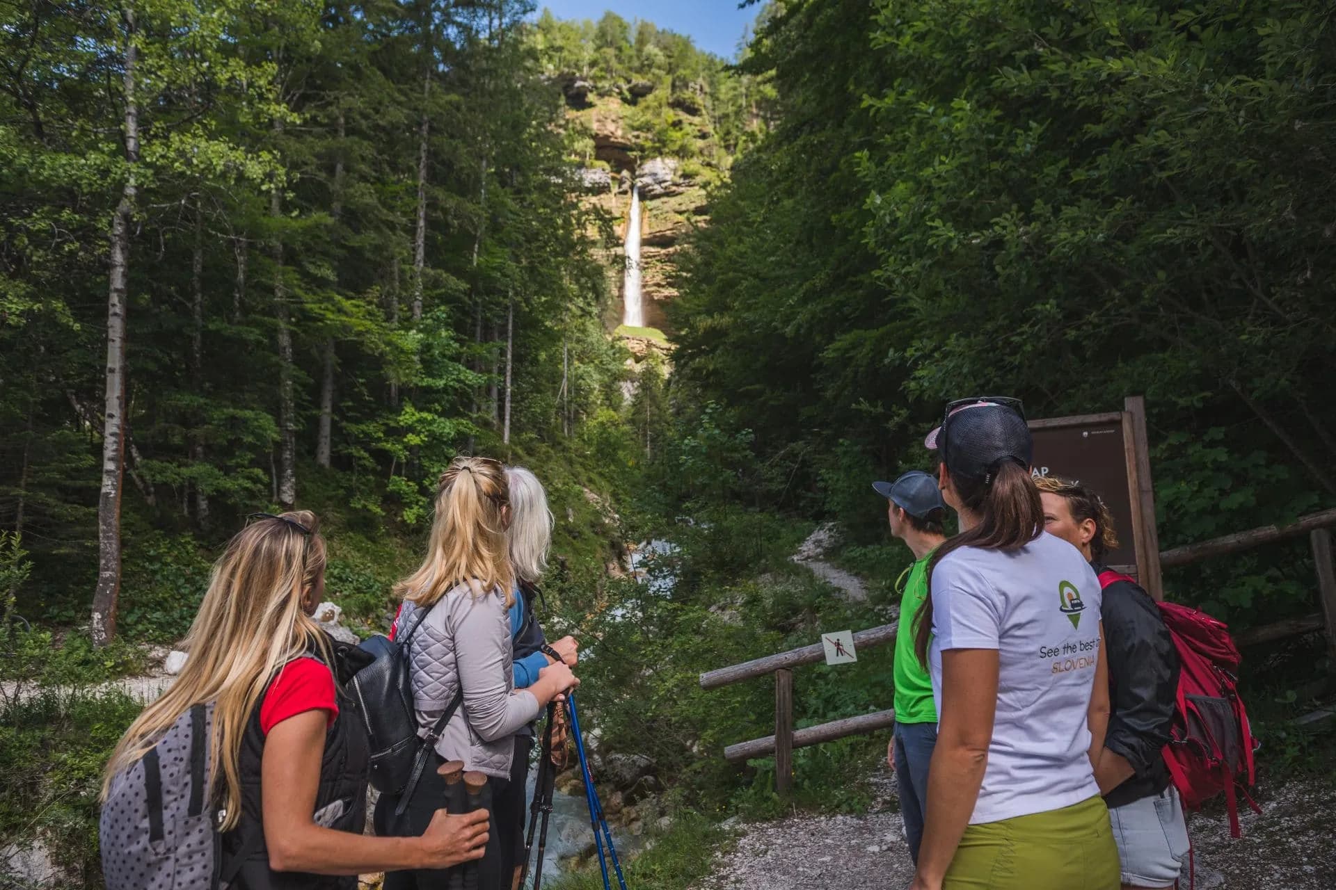 Hikers viewing Peričnik Waterfall cascading down a cliff in a dense green forest.